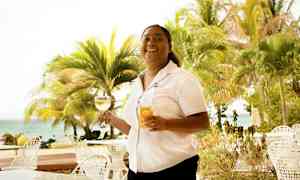 En person holder to glas, et med vin og et med juice, ved et bord udendørs nær stranden og omgivet af palmer. (AI-genereret tekst)