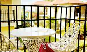 En altan med to hvide flettede stole og et rundt bord. På bordet ligger en bog. Udsigt over beplantning og bygninger i baggrunden. (AI-genereret tekst)