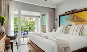 Hotelværelse med en hvid seng, dekorativ vægkunst med palmeblade, udsigt til en balkon med frodigt miljø. (AI-genereret tekst)