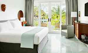 Et lyst og elegant hotelværelse med en stor seng, en lænestol og møbler med trædetaljer. Værelset har store glasdøre, der fører ud til en balkon med udsigt over frodige palmer. (AI-genereret tekst)