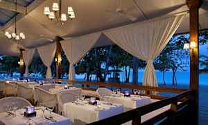 En restaurant med borde dækket med hvide duge ved stranden under et aftenbelyst telt. (AI-genereret tekst)