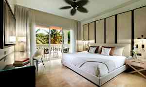 Et moderne soveværelse med stor seng, dekorativ vægpanel og åbning til en balkon med palmer udenfor. (AI-genereret tekst)