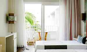 Lyst hotelværelse med to enkeltsenge og en altan med stole og bord. (AI-genereret tekst)