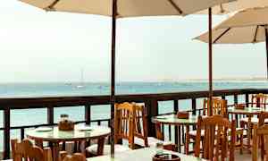 Udendørsborde og stole under parasoller med udsigt over havet. (AI-genereret tekst)