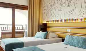 Et hotelværelse med to enkeltsenge med turkise dekorationer, bag sengene er der en væg med kunstnerisk dekoration og en lille lampe. (AI-genereret tekst)