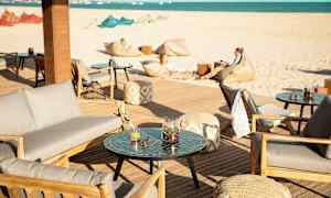 Terrasse med siddepladser og borde, i baggrunden en sandstrand og havet. (AI-genereret tekst)