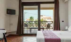 Et værelse med en seng, et lille TV på væggen og en balkon med udsigt over bygninger og palmer. (AI-genereret tekst)