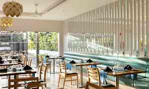 En moderne og lys restaurantindretning med borde og stole, hvor vinduer vender mod et frodigt udendørsområde. (AI-genereret tekst)