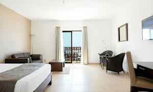 Et hotelværelse med lys indretning. En seng, sofa, bord og stole. En balkon med udsigt ses gennem glasdøre. (AI-genereret tekst)