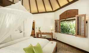 Et lyst værelse med et hvidt loft, en seng med myggenet og grønne puder, et skrivebord og en stol ved et vindue med udsigt over beplantning. (AI-genereret tekst)