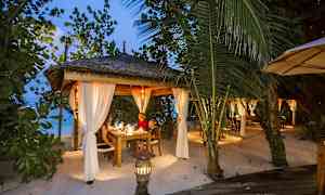 En oplyst udendørs terrasse med tag og hvide gardiner, omgivet af grønt på en strand. (AI-genereret tekst)