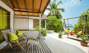 Et udendørsområde med træstole, en gynge og sand, omgivet af grønne planter og palmer. (AI-genereret tekst)