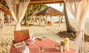 Borde dækket med servietter og tallerkener på sanden ved kysten, omgivet af tag med tekstildekorationer. (AI-genereret tekst)