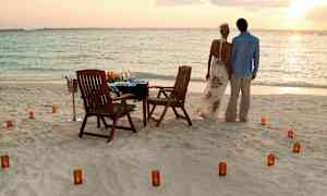 Et par står på en strand ved havet, foran et bord med stole og lys. (AI-genereret tekst)