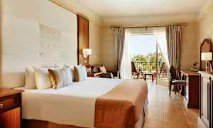 Et hotelværelse med stor seng, skrivebord og balkon med udsigt over grønt område. (AI-genereret tekst)