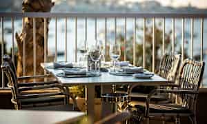 Et dækket bord med glas og servietter på en udendørs terrasse, omgivet af udsigt over vandet. (AI-genereret tekst)