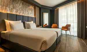 Et hotelværelse med to enkeltsenge, et stort vinduesparti med gardiner, to orange stole og et bord. (AI-genereret tekst)