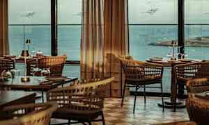 En restaurant med flettede stole og dækkede træborde med udsigt over havet gennem store vinduer. (AI-genereret tekst)