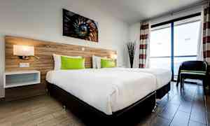 Hotelværelse med to enkeltsenge, hvidt sengetøj og grønne puder, et sengegærde af træpanel og store vinduer, der lukker naturligt lys ind. (AI-genereret tekst)