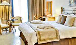 Et hotelværelse med en stor seng, sengetøj og puder. To stole og et bord står ved siden af, og en arbejdsplads med spejl og skrivebord ses i baggrunden. (AI-genereret tekst)