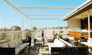 Billede af en tagterrasse med siddegrupper, borde og parasoller. Udsigt over bygninger og en klarblå himmel. (AI-genereret tekst)