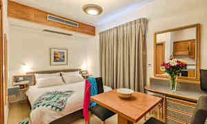 Et lyst hotelværelse med en dobbeltseng, spisebord, siddepladser og et spejl. Dekoreret med en blomsterkrukke på bordet. (AI-genereret tekst)