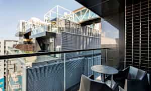 En balkon med moderne indretning, inklusive et rundt bord og stole, med udsigt over en bygning med glas- og metalfacader. (AI-genereret tekst)