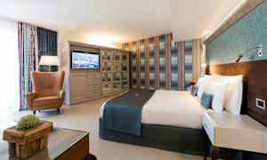 Et hotelværelse med en dobbeltseng, en lænestol, et bord og et tv på væggen. (AI-genereret tekst)