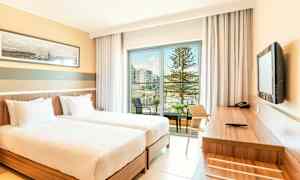 Et hotelværelse med to enkeltsenge, et skrivebord og udsigt til en balkon med pool og træer. (AI-genereret tekst)