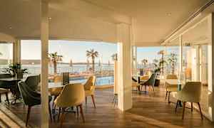 En restaurant med siddepladser og glasvægge med udsigt over en pool og palmer ved havet. (AI-genereret tekst)