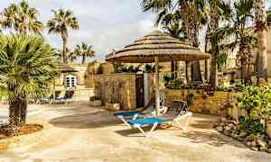 En stenbelagt terrasse med liggestole og halmtækte parasoller omgivet af palmer og stenbygninger. (AI-genereret tekst)