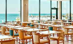 Restaurant med flere borde og stole, stor vindudsigt til havet. (AI-genereret tekst)