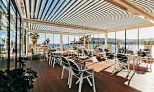 En udendørs terrasse med borde og stole, omgivet af glasvægge og med udsigt over havet og palmer. (AI-genereret tekst)