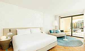 Et moderne hotelværelse med en stor seng, lyse vægge og elegant indretning. Værelset har et siddeområde med puder samt en rummelig balkon med udsigt. (AI-genereret tekst)