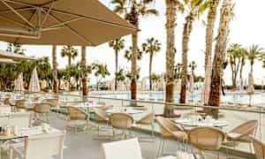 En udendørs restaurant med flere opstillede borde og stole, omgivet af palmer og med udsigt over en pool. (AI-genereret tekst)