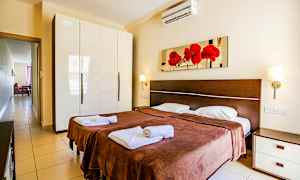 Et soveværelse med to enkeltsenge, brune sengetæpper og hvidt sengelinned. På væggen over sengene hænger et billede med røde blomster. Til venstre er der et garderobeskab med blank hvid overflade. (AI-genereret tekst)