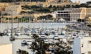 En marina med mange både, omgivet af historiske bygninger og frodig vegetation. (AI-genereret tekst)