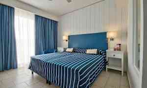 Et hotelværelse med en dobbeltseng med blå- og hvidstribede sengesæt, en blå sengeryg og hvide møbler. (AI-genereret tekst)