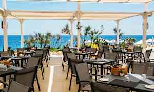 En terrasse med udendørsservering, borde og stole under et hvidt tag, med havet og vegetation i baggrunden. (AI-genereret tekst)