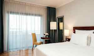 Et hotelværelse med en stor seng, et bord med en stol og et vindue dækket af hvide gardiner. (AI-genereret tekst)