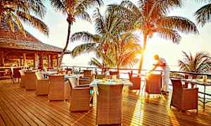 Udendørs serveringsområde med udsigt over havet og solnedgangen, palmer i baggrunden og en tjener, der forbereder et bord. (AI-genereret tekst)