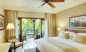 Et smagfuldt indrettet hotelværelse med en stor seng, siddeområde og en altan med udsigt over frodige palmer. (AI-genereret tekst)