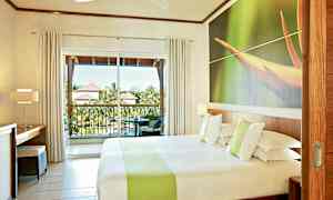 Hotelværelse med en dobbeltseng, en stor vinduesvæg og en balkon med udsigt over grønne områder og tage på nærliggende bygninger. (AI-genereret tekst)