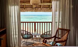 Udsigt over havet fra et værelse med en overdækket altan. Mellem gennemsigtige gardiner ses to havestole ved et bord. (AI-genereret tekst)