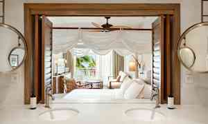 Et hotelværelse med siddeområde, seng og åbning mod en veranda. I forgrunden ses et badeværelse med dobbelt håndvask og spejl. (AI-genereret tekst)