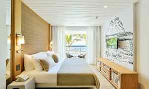 Et lyst hotelværelse med en dobbeltseng, vægmonteret TV og store vinduer med udsigt til havet. (AI-genereret tekst)