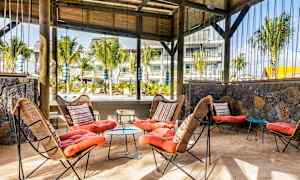 Et udendørsområde med siddepladser, med kurvestole og farverige puder samt et bord på sandgulvet. Trækonstruktion og grønne planter ses i baggrunden. (AI-genereret tekst)