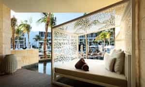 En liggestol med hvide puder, placeret udendørs omgivet af en hvid dekorativ struktur. I baggrunden ses palmer, en pool og moderne bygninger. (AI-genereret tekst)