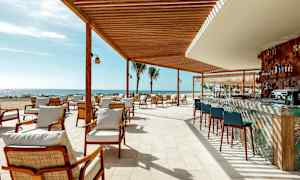 En udendørsrestaurant med siddepladser og en bar under en træpavillon ved stranden. (AI-genereret tekst)