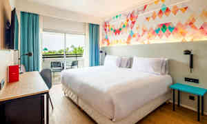 Et hotelværelse med en dobbeltseng, farverigt vægdesign, skrivebord og balkon med to stole. (AI-genereret tekst)